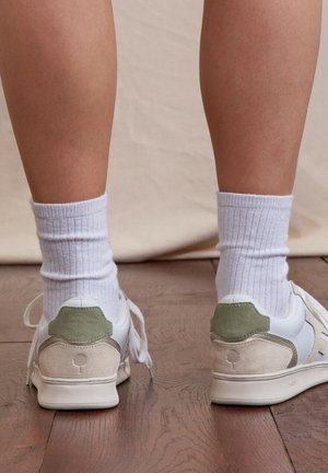 Person wearing white sneakers with green and beige accents and white ribbed crew socks, standing on wooden floor facing away.