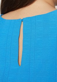 Blaues strukturiertes Gewebe mit einem quadratischen Muster, das eine Schlüsselaugenöffnung am Rücken aufweist, gesichert mit einer Schlaufe und einem Knopfdetail.