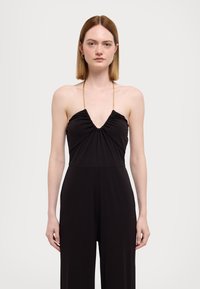 Salopette noire à encolure plongeante, avec un tissu froncé au niveau du corsage et une silhouette allongée avec des jambes larges. Bretelles avec accents dorés.