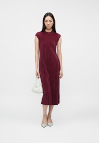 Γυναίκα που στέκεται, φορώντας ένα αμάνικο, ριμπ και μπορντό midi φόρεμα, κρατώντας μια μικρή λευκή τσάντα και φορώντας λευκά μυτερά παπούτσια.