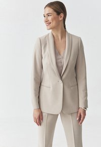 Beige puku na shingo ya shali, kufungwa kwa kitufe kimoja, na muundo ulioangaziwa. Kitambaa laini chenye mifuko ya pembeni na blouse inayolingana chini.