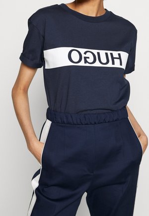 Navyblauwe katoenen t-shirt met opgerolde mouwen, voorzien van een witte horizontale band met vetgedrukte zwarte "HUGO" letters over de borst.