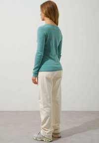 Maglione in cashmere turchese con maniche lunghe, design aderente; abbinato a pantaloni bianchi ampi e sneaker grigi. Sfondo semplice.