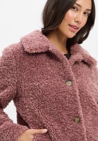 Frau trägt einen mauvefarbenen, strukturierten Teddy-Mantel mit großen Kragen, die Hand in der Tasche, schaut leicht zur Seite.