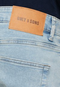 Vaaleansiniset farkut, joissa on nahkainen merkki vyötärönauhassa, johon on painettu tummilla kirjaimilla "ONLY & SONS".