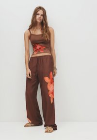 Giovane donna con lunghi capelli ondulati, che indossa un top senza maniche floreale marrone e pantaloni a gamba larga coordinati, in piedi davanti a una parete bianca.