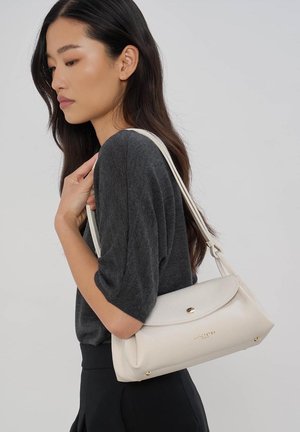 Vrouw met lang donker haar, een donkergrijze top en een zwarte broek, met een witte schoudertas met een klep en gouden details.