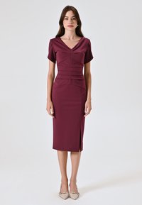 Burgunderfarbene taillierte Kleid mit kurzen Ärmeln, V-Ausschnitt, Raffung und seitlichem Schlitz, kombiniert mit hellbeigen Absatzschuhen.