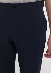 Tumedad sinised püksid, mis on valmistatud siledast kangast, omavad tasaset esiosa, ühte nööbi kinnitust ja on kohandatud disainiga vööde jaoks.