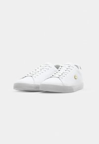 White sneakers with a smooth leather upper and a textured rubber sole. Features flat laces, gold logo accent, and minimalistic design.