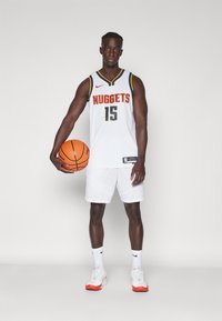 Basketbalista v bielom drese Denver Nuggets s žltými a červenými akcentmi, drží oranžovú basketbalovú loptu, má na sebe biele šortky a topánky.