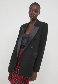 Frau trägt ein schwarzes Satin-Revers-Blazer über einem schwarzen Oberteil und einem rotkarierten Rock vor einem schlichten hellen Hintergrund.