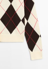 Manica di maglione con motivo a rombi argyle crema e marrone scuro, accentuata da sottili linee diagonali rosse e polsino a coste crema.