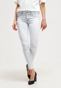 Žena v světle modrých skinny džínách s mírným popraskaním a námořnicky modrých lodičkách, stojící s překříženýma nohama.
