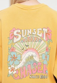 Gul genser med stort, fargerikt grafisk design. Elementene inkluderer bølger, blomster og tekst som sier "Sunset Roxy Chaser siden 1990."