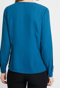 Bluzka z długim rękawem w kolorze morskim, z okrągłym dekoltem, gładką teksturą i mankietami na guziki; posiada prosty krój i minimalne zdobienia.