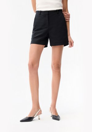 Kvinde iført skræddersyede sorte shorts og sort-hvide kitten heel slingback sko, stående mod en hvid baggrund.