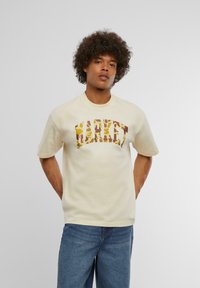 Cremefarvet t-shirt med korte ærmer og "MARKET" logo i et brunt og gult camouflage-mønster, lavet af blødt bomuldsstof.