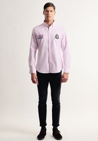 Camisa de botones rosa claro con cuello, que presenta texto bordado en azul "POLO LEGEND" y un diseño de insignia. Se lleva con jeans oscuros y zapatos.