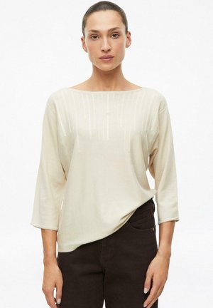 Frau mit zurückgebundenem dunklem Haar, die ein hellbeiges Dreiviertelarmshirt und dunkle Hosen trägt, steht vor einem weißen Hintergrund.