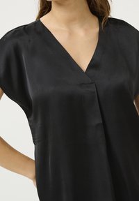 Femme portant un haut sans manches en satin noir à décolleté en V avec une texture lisse et brillante, la main posée sur la hanche.