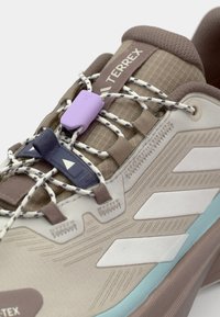 Lichtbruine buitenschoen met een gestructureerde bovenkant. Heeft een lichtgewicht ontwerp, elastische veters, paarse en blauwe accenten en witte branding.