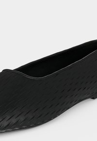 Μαύρο υφασμάτινο παπούτσι τύπου slip-on με μυτερή μύτη και χαμηλό τακούνι, πλάγια όψη που δείχνει την εξωτερική πλευρά και το άνοιγμα σε μονόχρωμο φόντο.