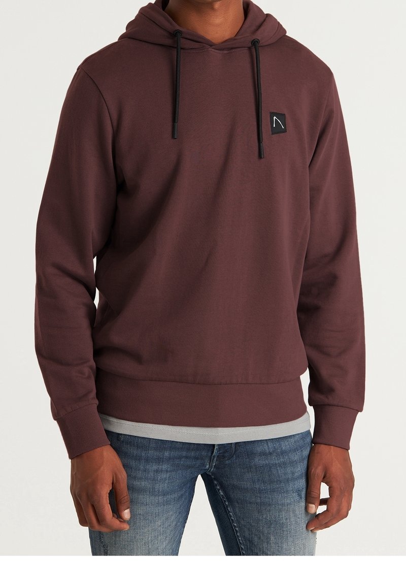 Homme portant un sweat à capuche marron avec des cordons noirs et un jean bleu sur un fond uni.