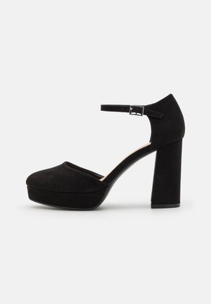 Chaussure à talon haut en daim noir avec talon épais, semelle plateforme, bout rond et bride à la cheville avec boucle argentée sur fond blanc.