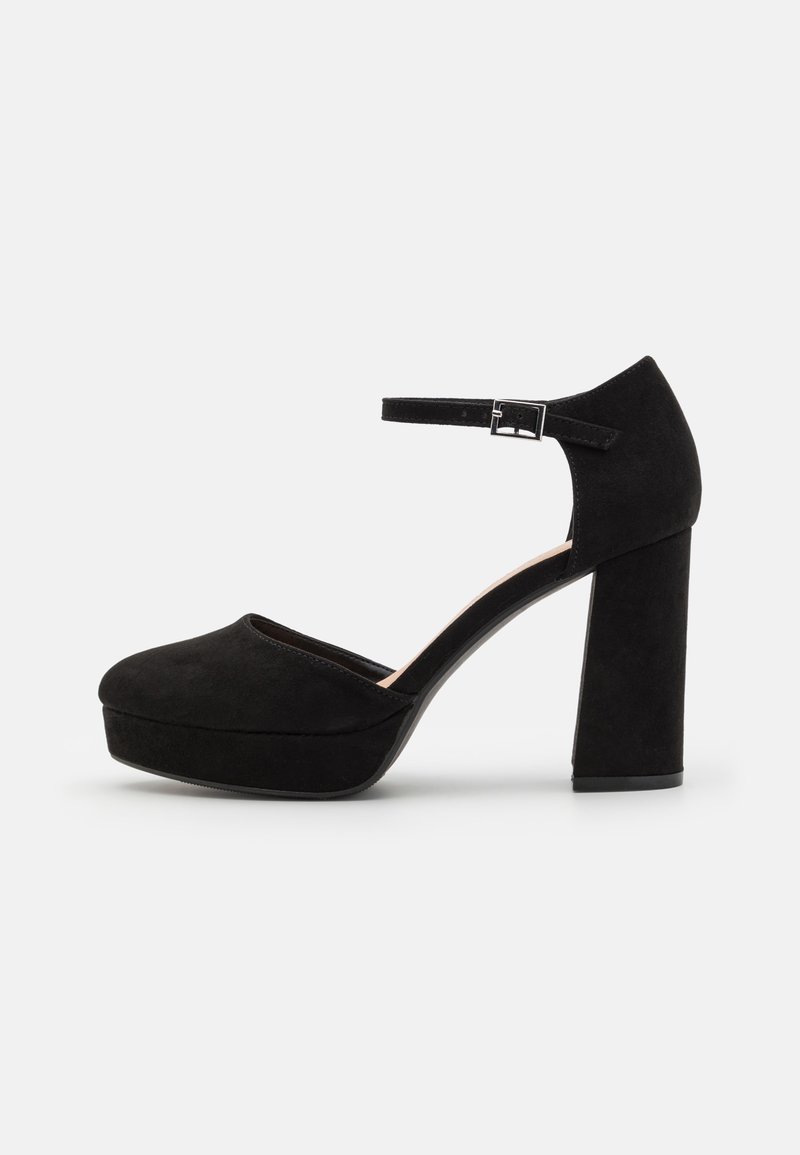 Chaussure à talon haut en daim noir avec talon épais, semelle plateforme, bout rond et bride à la cheville avec boucle argentée sur fond blanc.