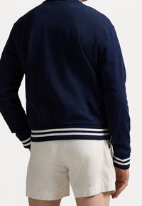 Mörkblå bomberjacka i bomull med vita randiga ribbade ärmmuddar och fåll, med en slät textur och avslappnad design.