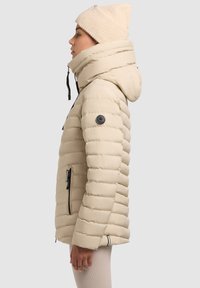 Béžová zateplená bunda s kapucňou, s horizontálnou prešívanou štruktúrou a čiernymi zipsovými vreckami. Nosená s svetlo béžovou pletenou čiapkou.