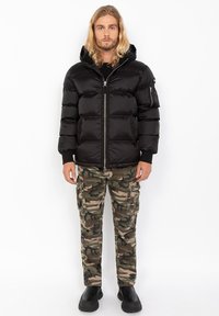 Sort pufferjakke med lynlås foran og hætte, der har en blank overflade, parret med camouflage cargobukser og sorte slip-on sko.