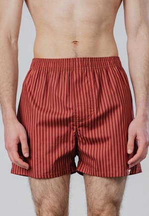 Erwachsener Mann trägt rot und dunkelrot gestreifte Shorts mit elastischem Bund, steht mit locker hängenden Armen an den Seiten.