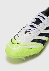 Botas de fútbol en blanco, negro y verde neón con un upper sintético texturizado, rayas contrastantes y tacos transparentes en la suela. Marca "HYBRIDTOUCH" visible.