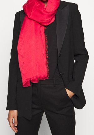 Personne portant un costume noir sur mesure avec la main dans la poche, associée à une grande écharpe rouge vif enroulée autour du cou.