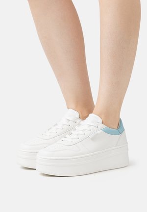 Sneakers bianchi con accenti azzurri indossati a piedi nudi, mostrati da un lato su uno sfondo bianco semplice.