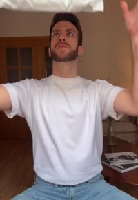 Weißes Kurzarm-T-Shirt an einem bärtigen Mann, der sich nach oben streckt. Teilweise sichtbare Holzmöbel und Zeitschriften im Hintergrund.