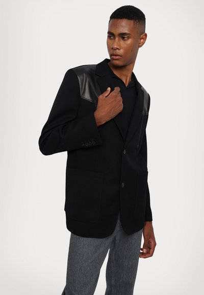 Zwarte blazer met leren accenten op de schouders, twee voorzakken, sluiting met één knoop en een getailleerde pasvorm, gecombineerd met grijze jeans.