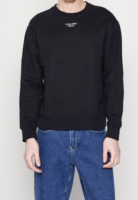 Hombre vistiendo un sudadera negra de Calvin Klein Jeans con texto blanco del logo en el pecho y jeans de mezclilla azules, de pie frente a un fondo blanco liso.