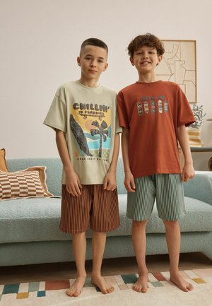 Zwei barfüßige Jungen stehen drinnen, tragen bedruckte T-Shirts und gestreifte Shorts in einem Wohnzimmer mit Sofa und gerahmten Kunstwerken.