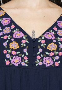Robe bleu marine avec broderies florales roses, violettes et jaunes ainsi que des boutons recouverts de tissu le long du col en V, dévoilant un petit tatouage sur la poitrine.