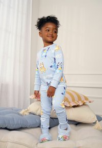 Pigiami di cotone blu con motivi di cani dei cartoni animati nei colori blu, arancione e giallo, con polsini a coste. Modello che indossa pantofole comode.