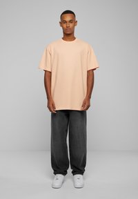 Pudderfarget oversized T-skjorte med korte ermer, kombinert med mørkegrå vide bukser og hvite joggesko, satt mot en grå bakgrunn.