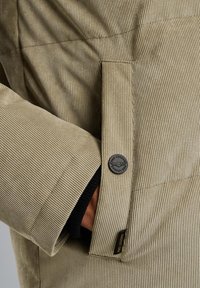 Beige corduroy-takki, jossa on tumma nappi ja mustat ribbit, näkyvät raidat ja taskudetaili sivussa.