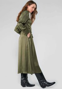 Robe en satin vert olive avec des manches longues et des poches latérales, présentant une texture lisse et un design fluide, associée à des bottes de cow-boy en cuir noir.