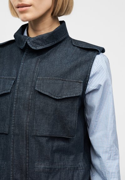 Γυναίκα που φοράει σκούρο τζιν αμάνικο γιλέκο με θήκες με καπάκια πάνω από ανοιχτό μπλε και λευκό ριγέ μακρυμάνικο πουκάμισο.