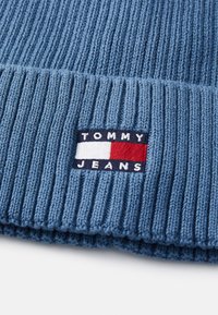 Plava rebrasta pletena kapa s logotipom koji prikazuje "TOMMY JEANS" u bijeloj i tamnoplavoj boji, uz crvene i bijele akcentue na plavoj pozadini.