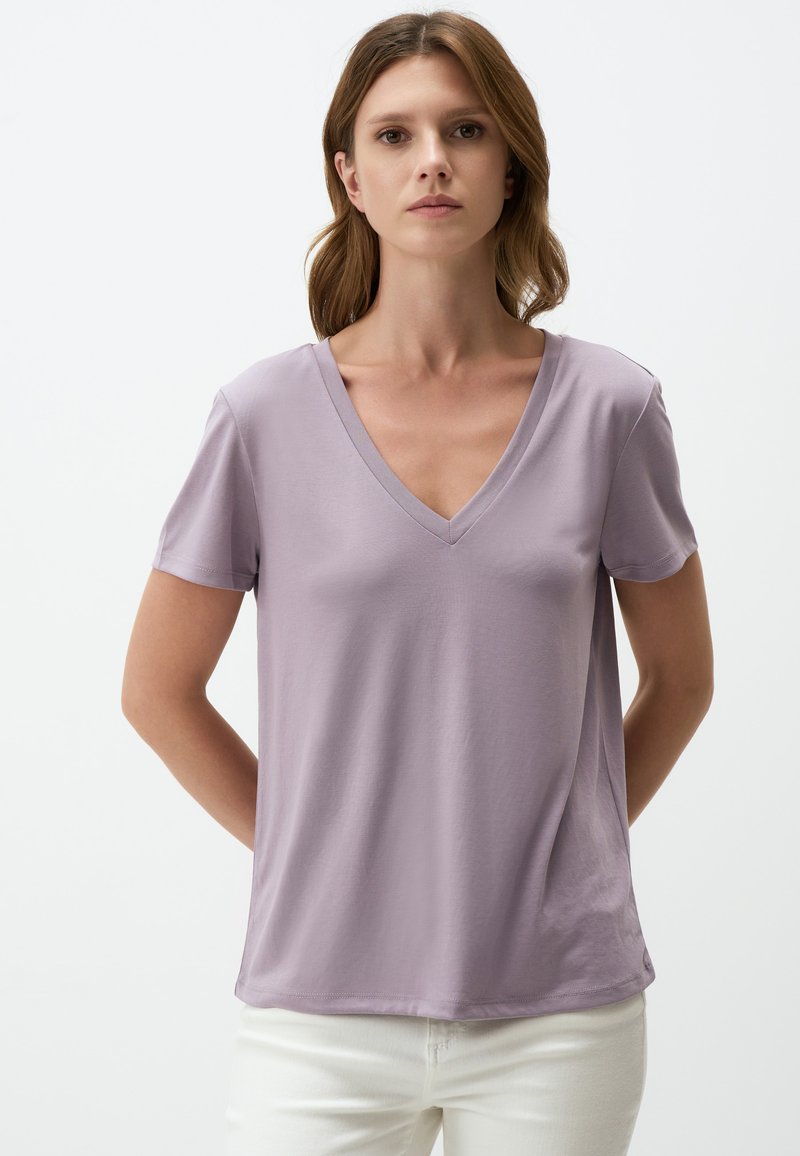 T-shirt lavanda a scollo a V realizzata in tessuto morbido con maniche corte, caratterizzata da una vestibilità rilassata e una texture liscia, abbinata a pantaloni bianchi.