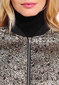 Metaliczna kurtka bomberka na suwak z wzorem przypominającym łuski w kolorach złotym, brązowym i czarnym. Pod spodem widoczna jest czarna dzianina z golfem.